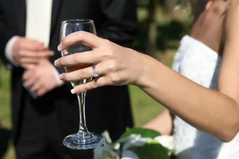 Dłoń panny młodej z pierścionkiem trzyma kieliszek szampana. W tle pan młody w garniturze. 38 rocznica ślubu nazwa.