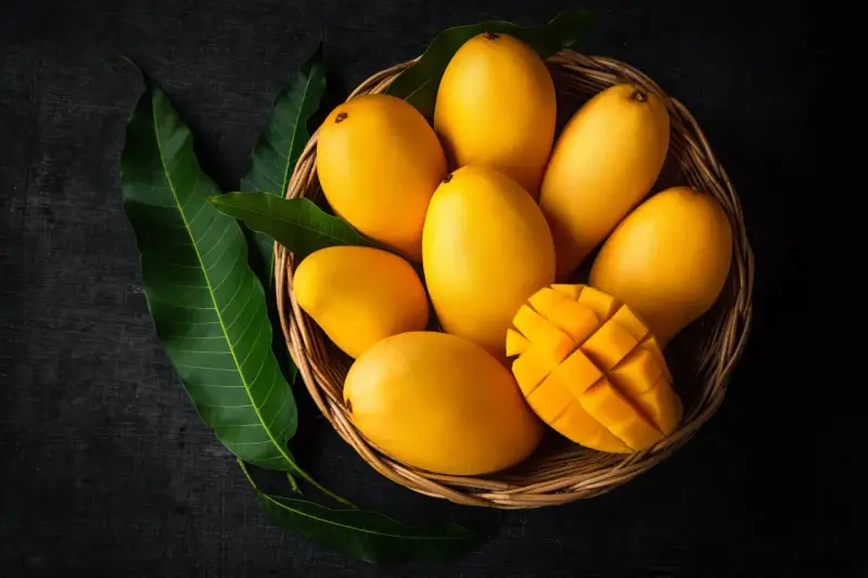 Koszyk pełen soczystych, żółtych mango, jedno przekrojone w kostkę. Obok zielone liście mango.