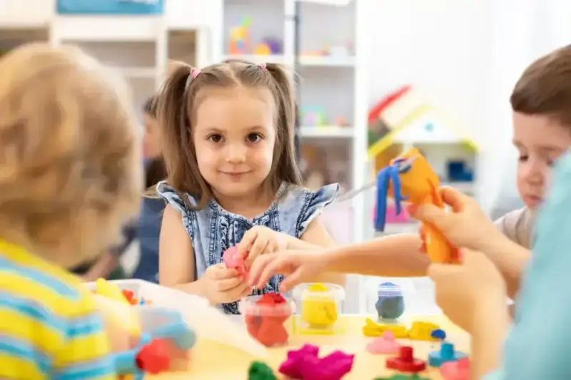 Uśmiechnięta dziewczynka z kitkami bawi się plasteliną, zastanawiając się, w jakim wieku do szkoły pójdzie. Dzieci tworzą kolorowe figurki.