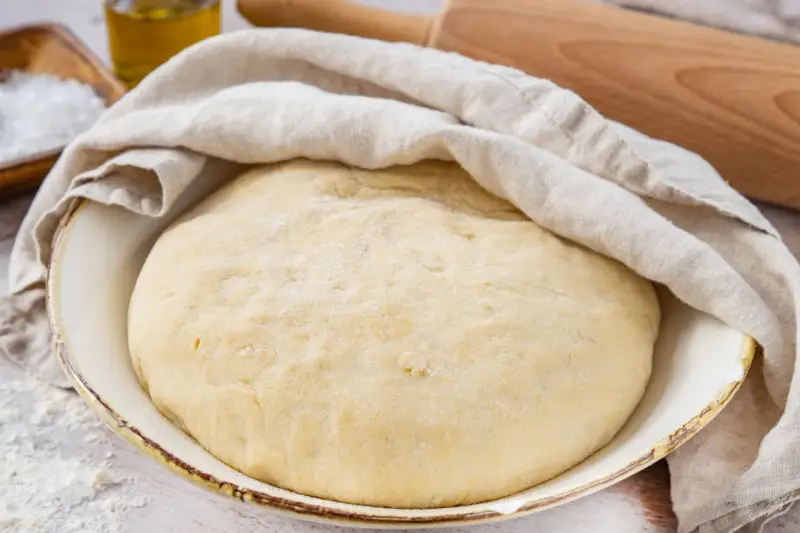 Der weltbeste Pizzateig mit Trockenhefe ruht abgedeckt in einer Schüssel. Daneben liegen Salz, Olivenöl und ein Nudelholz.