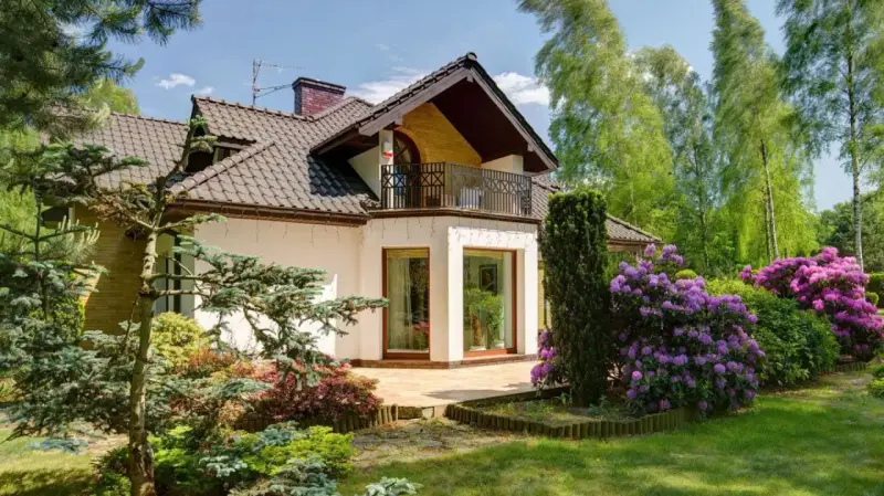 Dom z balkonem i tarasem, otoczony bujną zielenią i kwitnącymi rododendronami. Występ ściany podkreśla nowoczesny charakter budynku.