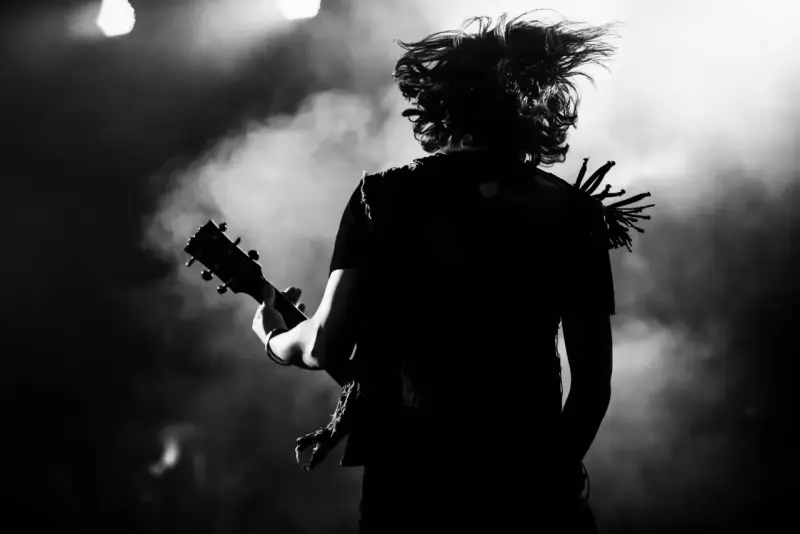 Muzyk z rozwianymi włosami gra na gitarze, w dymie i świetle sceny. Energia gatunku rockowego w czerni i bieli.