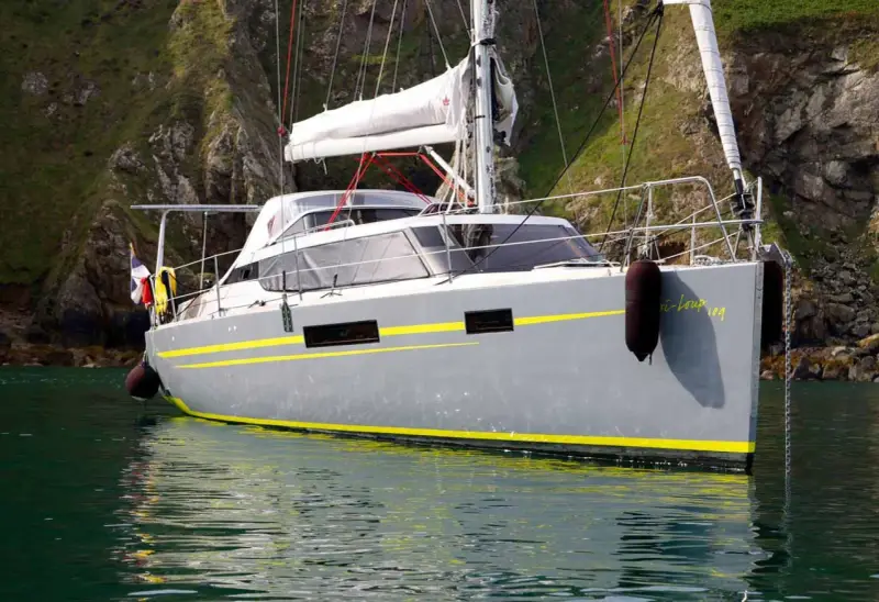 Le voilier "Bi-loup 109" gris avec des bandes jaunes, ancré près d'une falaise verdoyante.