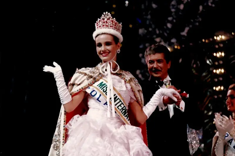 Uśmiechnięta Miss International w koronie i białej sukni, z szarfą "Miss Polonia". Obok niej mężczyzna w smokingu.