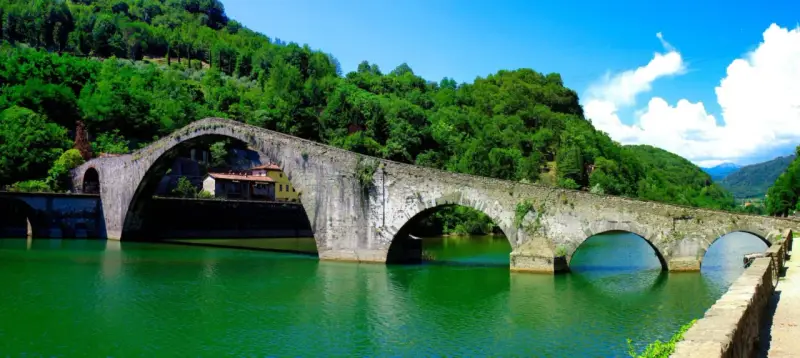 Il Ponte del Diavolo, con i suoi archi in pietra, attraversa un fiume verde smeraldo, circondato da colline lussureggianti e un cielo azzurro.
