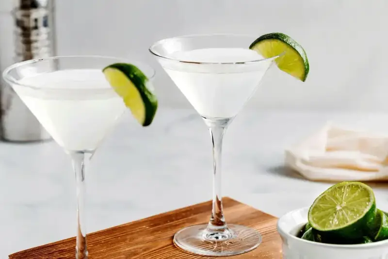 Dwa kieliszki z białym drinkiem, ozdobione plasterkami limonki. Obok miseczka z limonkami i shaker. Idealny kamikaze shot na gorący wieczór.