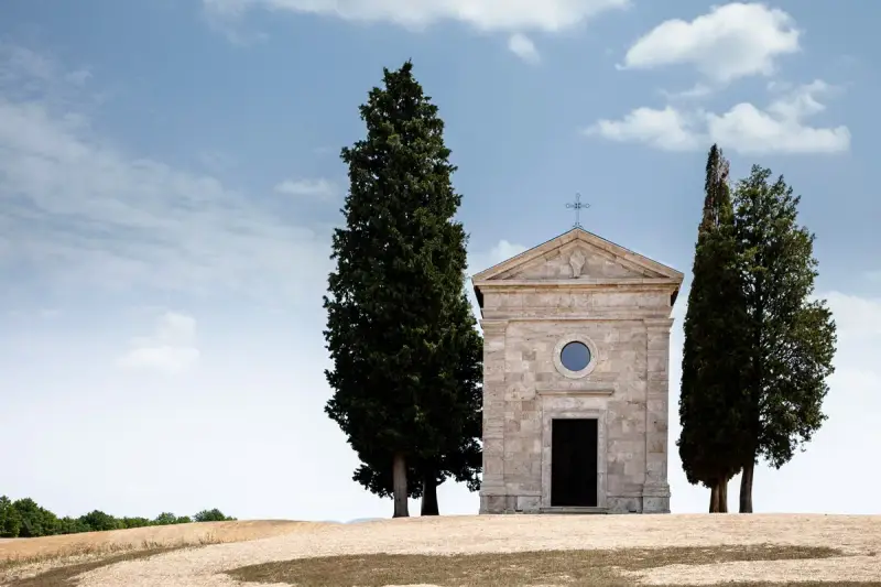 La cappella della Madonna di Vitaleta, con i suoi cipressi iconici, si erge solitaria su una collina toscana sotto un cielo azzurro e nuvoloso.