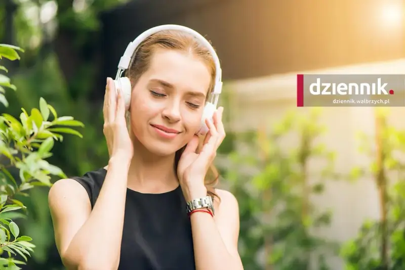 Kobieta w słuchawkach na uszach, z zamkniętymi oczami, cieszy się chwilą słuchania muzyki w otoczeniu zieleni.