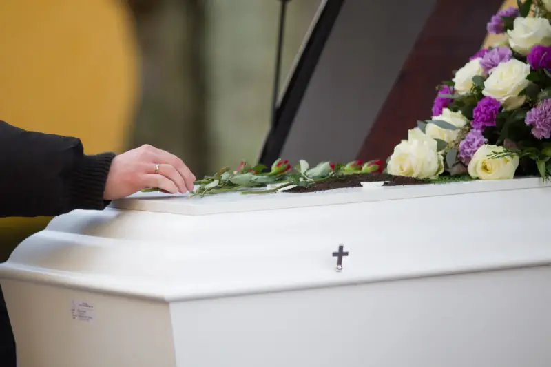 Dłoń z obrączką kładzie róże na białej trumnie, ozdobionej bukietem kwiatów i krzyżem.