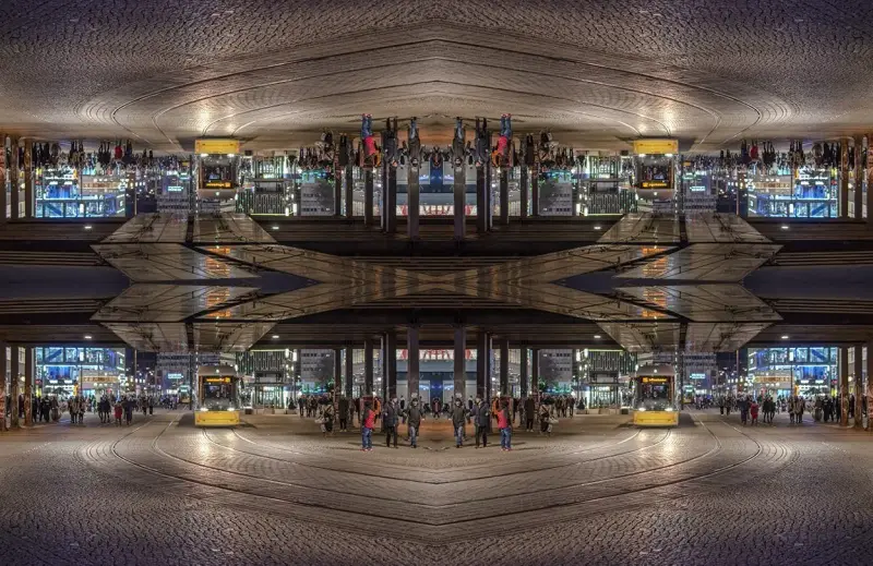 Nocne miasto z żółtym tramwajem i ludźmi. Kompozycja symetryczna tworzy surrealistyczny, odbity świat.