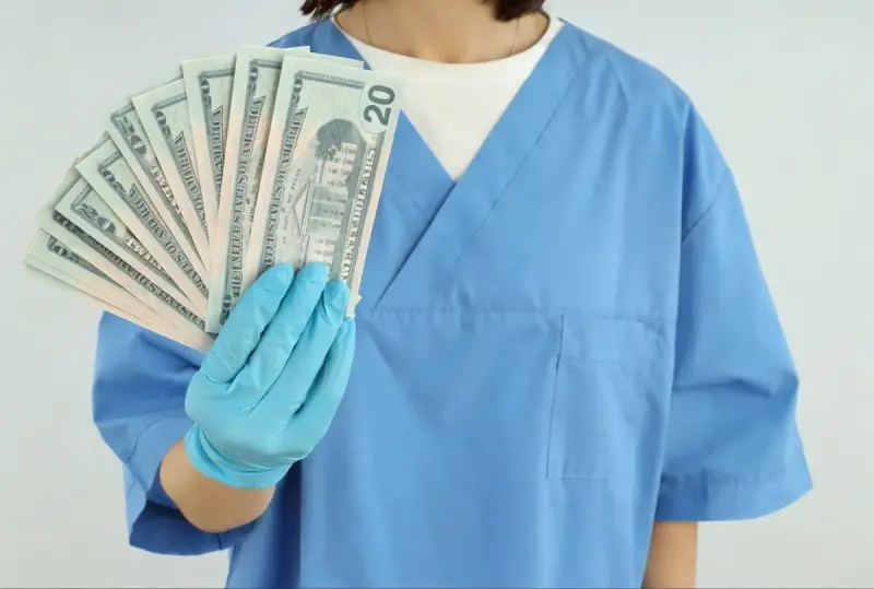 Higienistka stomatologiczna w niebieskim fartuchu pokazuje wachlarz banknotów 20 dolarowych. Zarobki netto mogą być satysfakcjonujące.