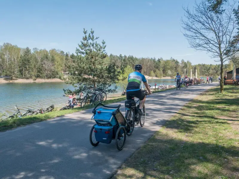 Rodzina na rowerach z przyczepką nad jeziorem Świerklaniec. W tle inni rowerzyści i plażowicze.