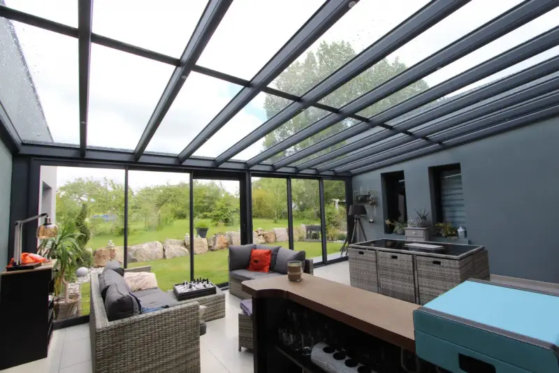 Salon moderne avec un toit en verre laissant entrer la lumière. Des canapés confortables invitent à la détente, tandis que le jardin verdoyant offre une vue paisible.