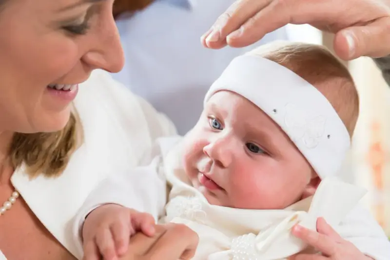 Uśmiechnięta mama trzyma dziecko w białej szatce. Dłoń ojca błogosławi malucha. Zastanawiasz się, ile kosztuje chrzest?