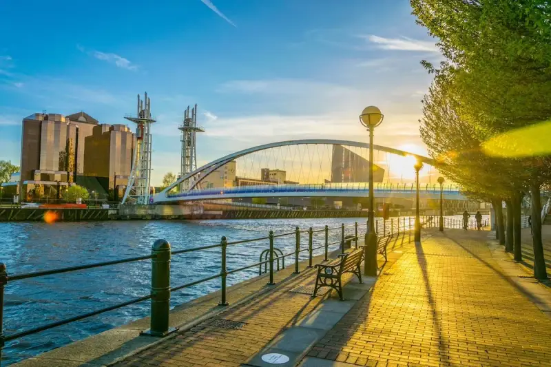 Promenade ensoleillée au bord de l'eau, avec un pont moderne et des bâtiments reflétant le soleil. Idéal pour visiter Manchester ou Liverpool.