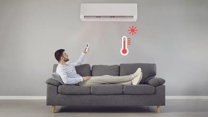 Hombre en sofá con control remoto ajustando el aire acondicionado calefacción. Termómetro rojo indica alta temperatura.