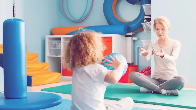 Ergotherapie für Kinder: Eine Therapeutin erklärt einem Kind mit Locken, wie man einen Ball wirft.