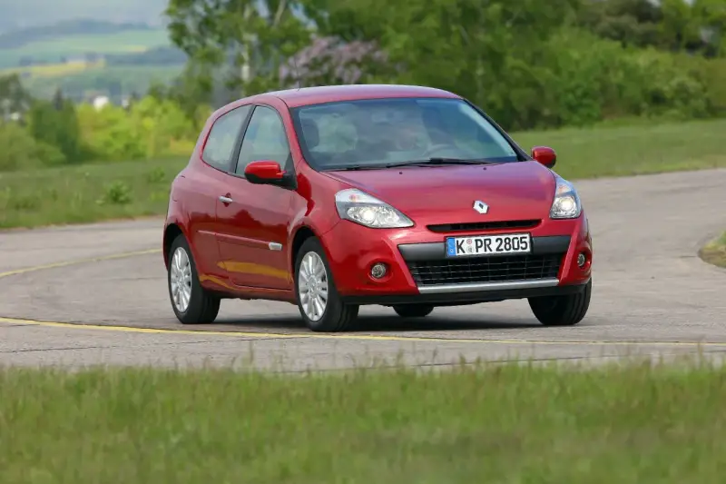 Czerwony Clio 3 na zakręcie drogi, otoczony zielenią.