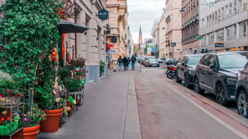 Wjazd do Wiednia samochodem osobowym. Ulica z kawiarniami, sklepami i zielenią, z zaparkowanymi autami i przechodniami.