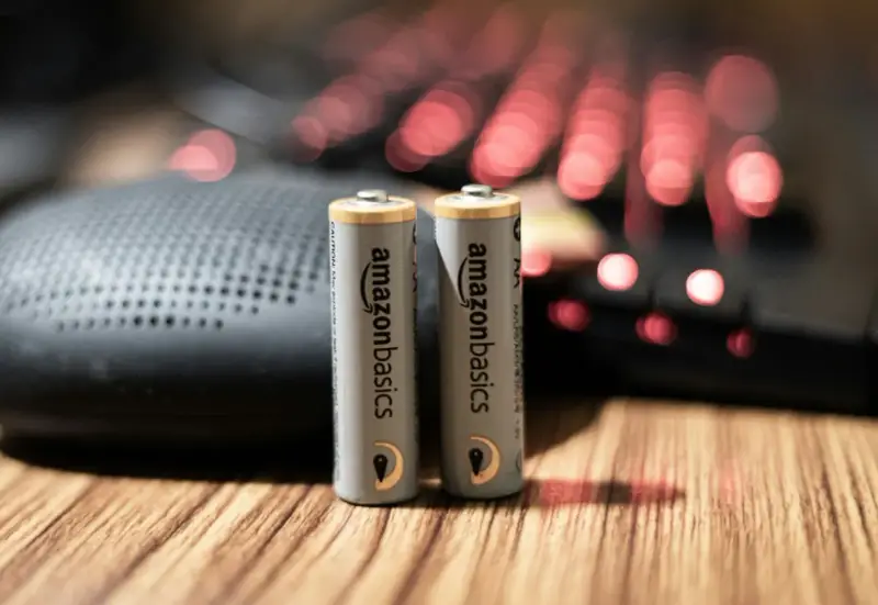 Dwie baterie paluszki AA AmazonBasics stoją na drewnianym blacie, obok głośnika i podświetlanej klawiatury.