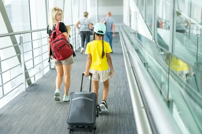 Młoda kobieta z plecakiem i dziewczynka z walizką idą korytarzem lotniska. To może być ich wyjazd nieletniego za granicę bez rodziców.