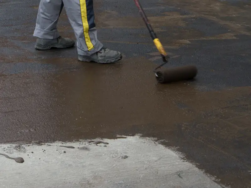 Pracownik wykonuje hydroizolację chudziaka, nakładając masę bitumiczną wałkiem.