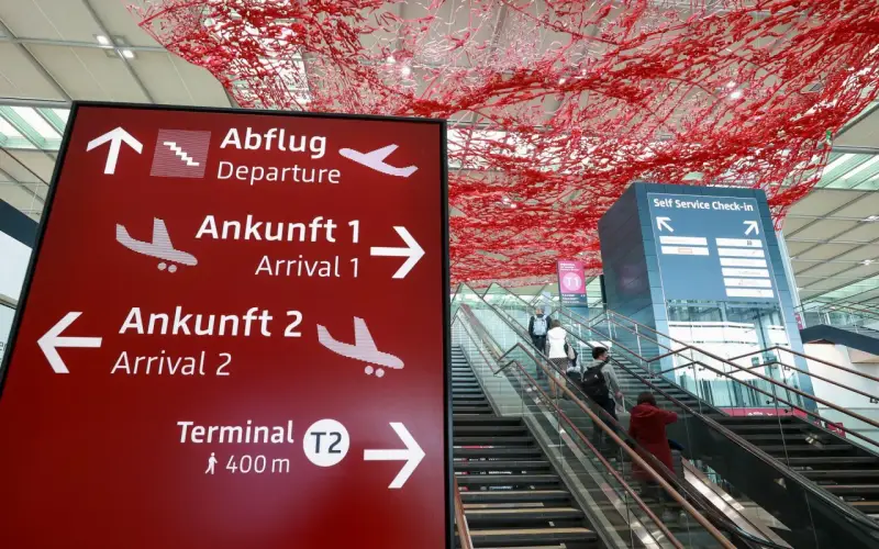 Czerwony znak z kierunkami na lotnisku w Niemczech: odloty, przyloty, terminal T2. W tle schody ruchome i instalacja artystyczna.