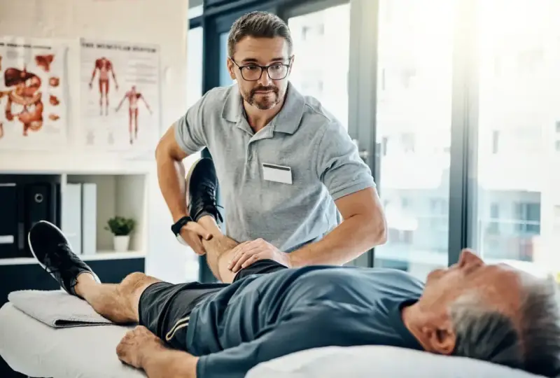Fizjoterapeuta ćwiczy nogę starszego pacjenta. Rehabilitacja jest kluczowa, by dowiedzieć się, komu przysługuje szpital uzdrowiskowy.
