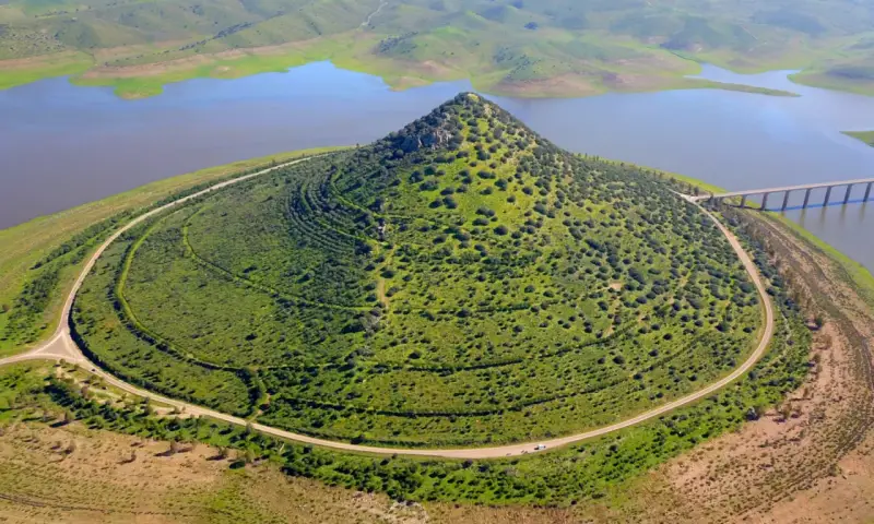 El **cerro Masatrigo**, cubierto de vegetación, se alza majestuoso junto a un embalse. Una carretera serpentea a su alrededor, conectando con un puente.