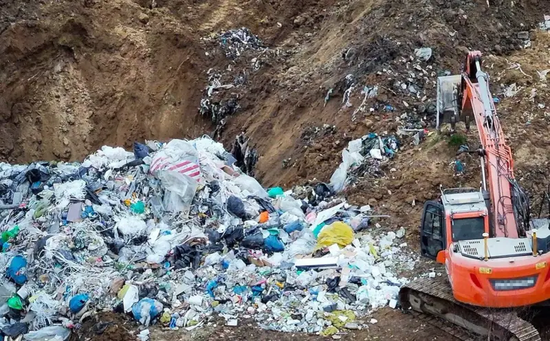 Ogromna góra śmieci, tworząca nielegalne wysypisko, z koparką w tle. Widać worki, folie i różne odpady.