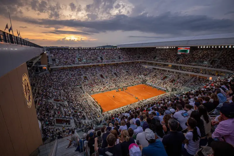 Widok na pełny stadion podczas meczu na kortach ziemnych. Zachodzące słońce oświetla trybuny pełne widzów, tworząc atmosferę emocjonującego **Roland Garros live**.