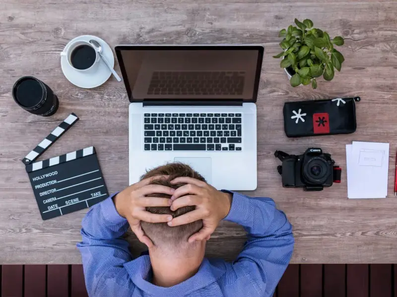 Frustracja twórcy filmowego. Na drewnianym biurku leży laptop, aparat, klaps, kawa i roślina.