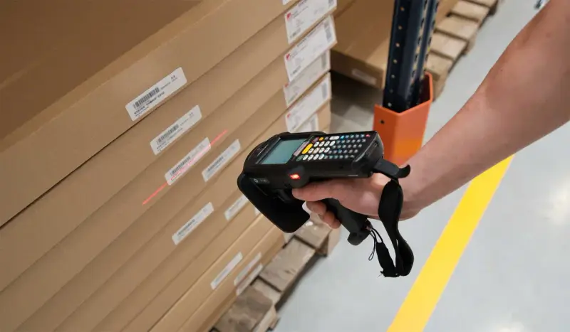 Mano escaneando cajas apiladas con un dispositivo, usando la **fórmula de Wilson** para optimizar el inventario.