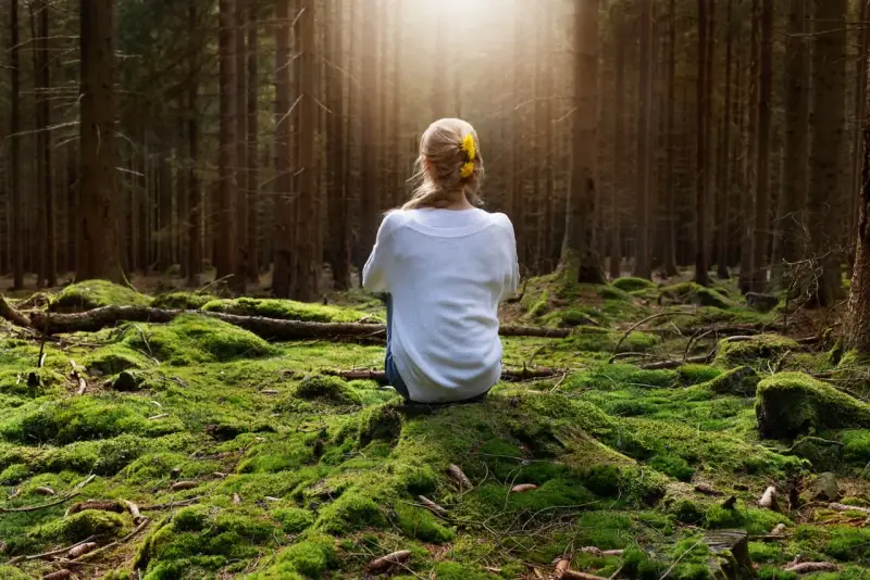 Frau sitzt im Wald auf moosbewachsenem Boden, umgeben von Bäumen. Sonnenstrahlen fallen durch das Blätterdach und erzeugen eine Atmosphäre der Katharsis.