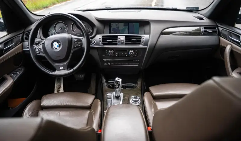 Wnętrze BMW X3 z brązową skórzaną tapicerką i drewnianymi wstawkami. Kierownica, deska rozdzielcza i nawigacja. Wiele osób szuka bmw x3 opinie, by poznać ten model.