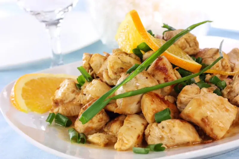 Sabroso pollo a la naranja, trozos jugosos con rodajas de naranja fresca y cebolleta picada, servido en un plato blanco.