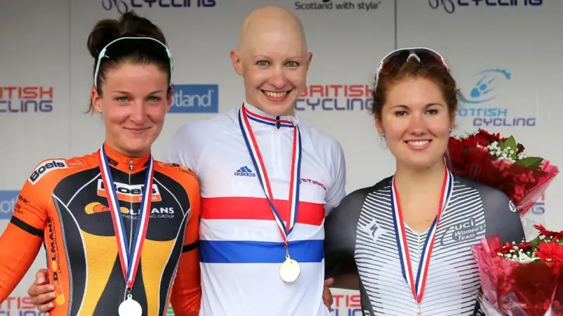 Joanna Rowsell, a bald cyclist in a white and red jersey, smiles on the podium with two other medalists.