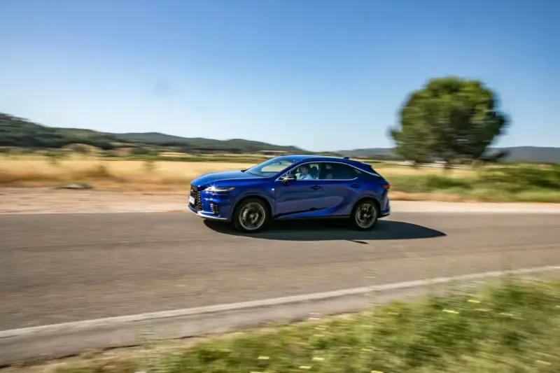Niebieski Lexus 450h pędzi po wiejskiej drodze, otoczony zielenią i wzgórzami.