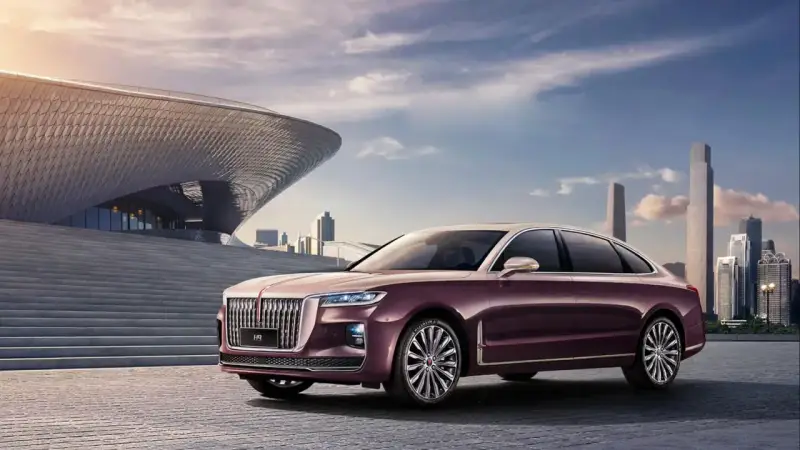 Elegancki, bordowy Hongqi H9 z chromowanymi detalami stoi przed nowoczesnym budynkiem o falistej fasadzie, w tle miejski krajobraz.