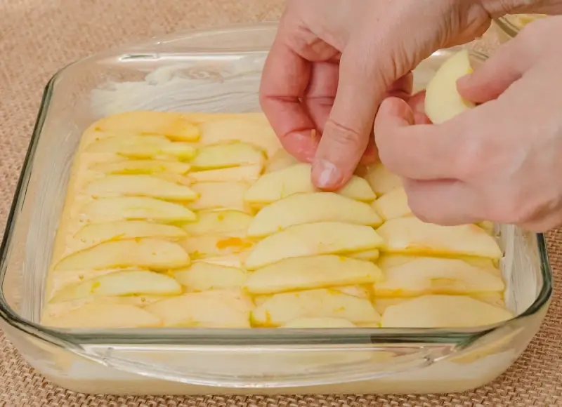 Ręce układają plasterki jabłek na cieście. Powstaje pyszne ciasto z jabłkami, gotowe do pieczenia.