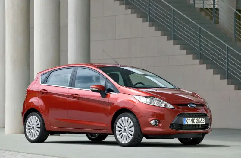 Czerwony Ford Fiesta MK6 zaparkowany przed nowoczesnym budynkiem z betonowymi kolumnami i schodami.