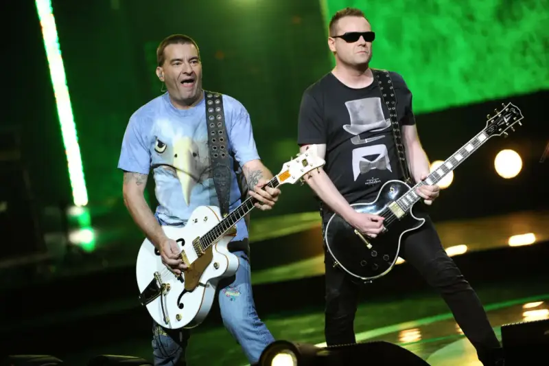 Dwóch członków Kobranocki gra na gitarach podczas koncertu. Jeden w koszulce z orłem, drugi w czarnej z cylindrem i muszką.