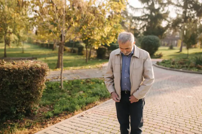 Starszy mężczyzna w beżowej kurtce Harrington i niebieskiej koszuli spaceruje po parku jesienią.