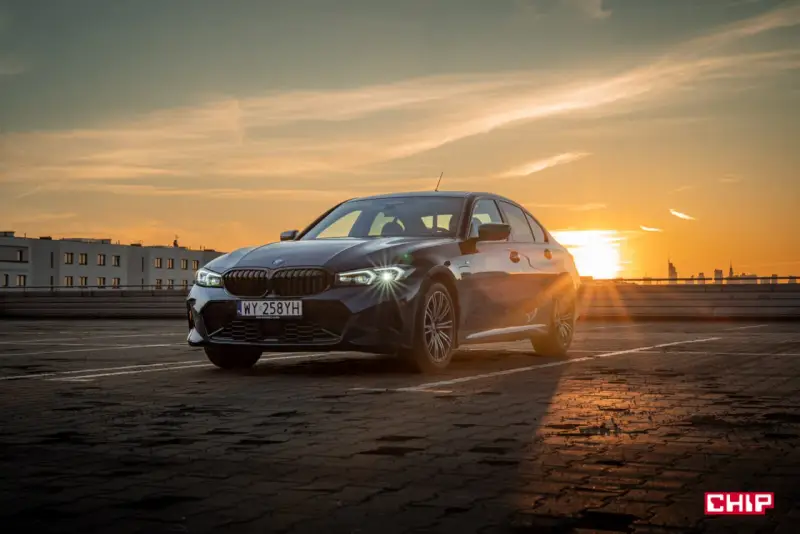 Ciemnoszara limuzyna BMW 330e na parkingu o zachodzie słońca.