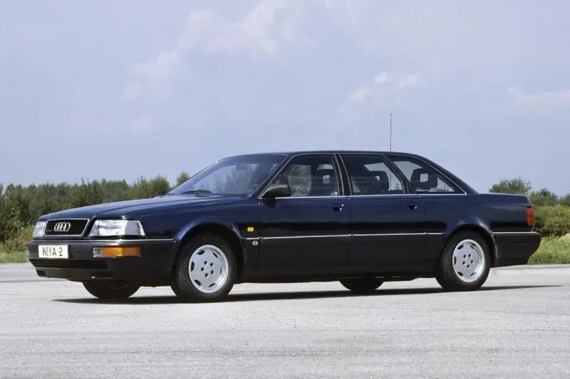 Ciemnoniebieski Audi V8 z lat 90. stoi na pustej drodze, z drzewami w tle.