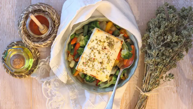 Feta al forno con verdure miste, condita con erbe aromatiche. Accanto, olio, spezie e un mazzetto di origano secco.