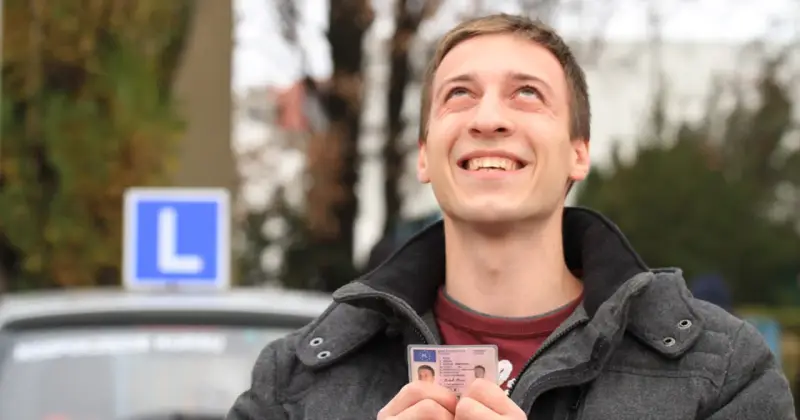 Szczęśliwy młody mężczyzna trzyma prawo jazdy kategorii B, z niebieskim znakiem "L" na dachu samochodu w tle.
