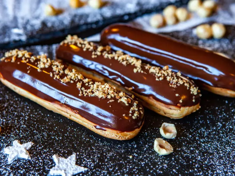Trois éclairs au chocolat brillants, saupoudrés de noisettes concassées, sur un fond sombre parsemé de sucre glace.