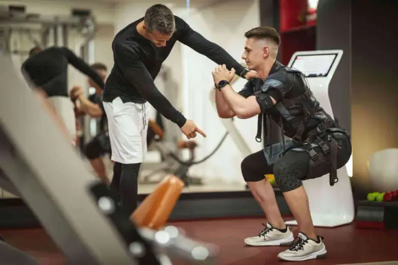 Trening EMS: Instruktor koryguje pozycję mężczyzny w kamizelce stymulującej mięśnie podczas przysiadu.