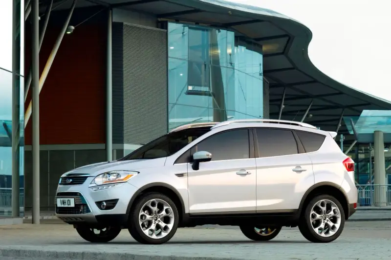 Biały Ford Kuga 2010 zaparkowany przed nowoczesnym budynkiem z przeszkloną fasadą.